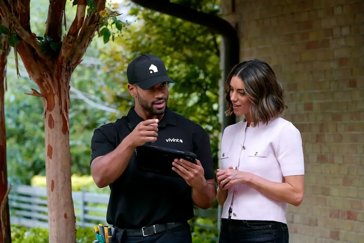 A Vivint service technician wearing a black uniform and hat shows information on a tablet to a female customer standing outside a brick home.